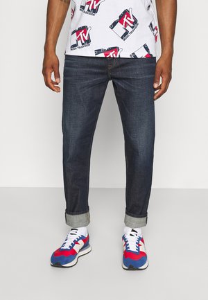 Homme portant un jean bleu foncé retroussé, des chaussettes blanches, des baskets colorées rouge, bleu et blanc, et un t-shirt blanc avec le logo MTV.