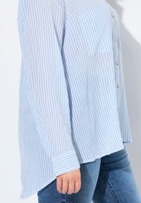 Gestreiftes blaues Hemd mit Knopfleiste aus leichtem Stoff, ausgestattet mit einer Brusttasche und einem abgerundeten Saum, kombiniert mit dunkelblauen Jeans.