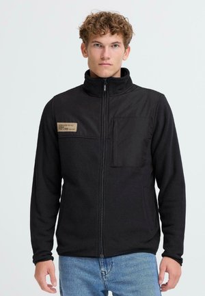 Black fleece jacket with a zip front, high collar, and a chest pocket. Features a textured panel and a small fabric tag with writing.