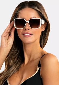 Duże kwadratowe okulary przeciwsłoneczne z białą oprawką i gradientowymi brązowymi soczewkami. Oprawka ma błyszczące wykończenie i subtelne akcenty metalowe.