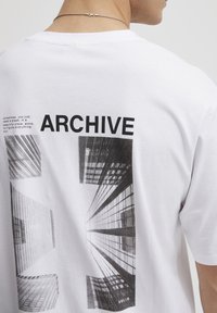 Hvid bomulds t-shirt med et grafisk tryk af skyskrabere, med teksten "ARCHIVE" i fed sort ovenfor billedet og "SOLID" nedenfor.