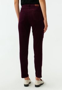 Pantalon en velours côtelé bordeaux à coupe ajustée, avec deux poches arrière et un détail en cuir à la taille. Texture lisse avec des nervures subtiles.