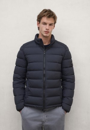 Young man wearing a black padded jacket with zip and gray pants, standing with hands in pockets against a plain wall.