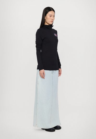 Black long-sleeve zip-up top with a small colorful graphic. Paired with a light blue denim maxi skirt and black shoes. Simple design, fitted silhouette.