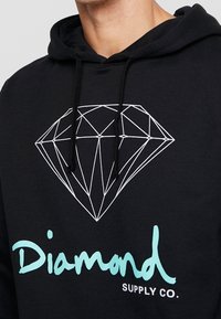 Svart hoodie med vitt geometriskt diamantgrafik och turkosa "Diamond"-text ovanför mindre vit "SUPPLY CO."-text på bröstet.