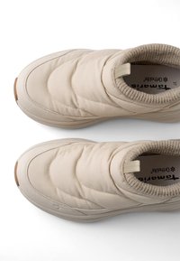 Ζευγάρι μπεζ παπούτσια slip-on με επένδυση, μαλακή υφασμάτινη επένδυση, στρογγυλεμένα δάχτυλα και ταινίες έλξης στους αστραγάλους σε λευκό φόντο.