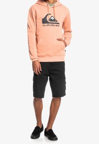 Koralli huppari mustalla logolla, etutasku ja säätönyöri huppu, yhdistetty mustiin cargo-shortsseihin ja mustiin liukusandaaleihin.