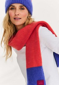 Czerwono-niebieski szalik w paski wykonany z miękkiego materiału, z prążkowaną fakturą na jednym końcu i jednolitym kolorem na drugim, oznaczony etykietą na dole.