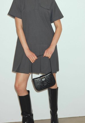 Rochie gri cu mânecă scurtă, cu un buzunar, design plisat, asortată cu o geantă neagră din piele ecologică cu imprimeu de crocodil și cizme lucioase negre până la genunchi.