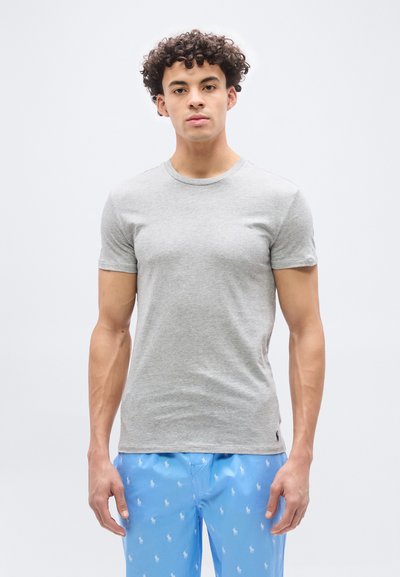 Νεαρός με σγουρά μαλλιά, ντυμένος με απλό γκρι μπλουζάκι και ανοιχτό μπλε σορτς με μικρά λευκά λογότυπα, στέκεται μπροστά σε απλό φόντο.