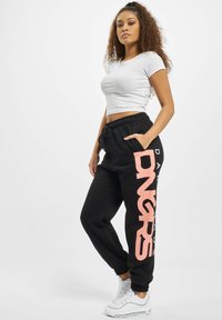 Black sweatpants with a large pink "DNGRS" logo, elastic waistband, side pockets, paired with a fitted white cropped t-shirt and white sneakers.