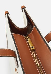 White canvas handbag with brown leather accents, featuring an open top, zipper closure, and contrasting stitching detail.