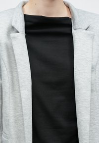 Világosszürke zakó strukturált gallérral, fekete, sima, rövid ujjú felső fölött viselve. A szövet finom textúrájú, látható minták nélkül.