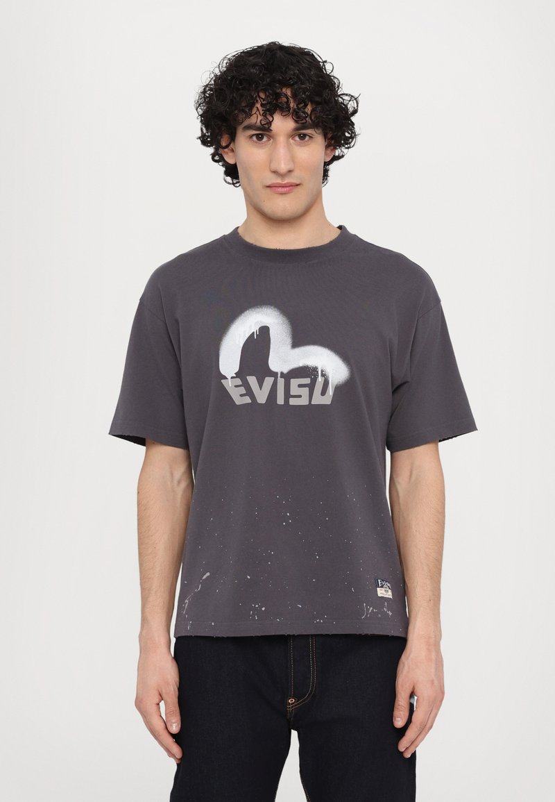 Camiseta gris de manga corta con cuello redondo, que presenta un logo pintado y detalles de salpicaduras en la parte frontal, hecha de tejido de algodón.