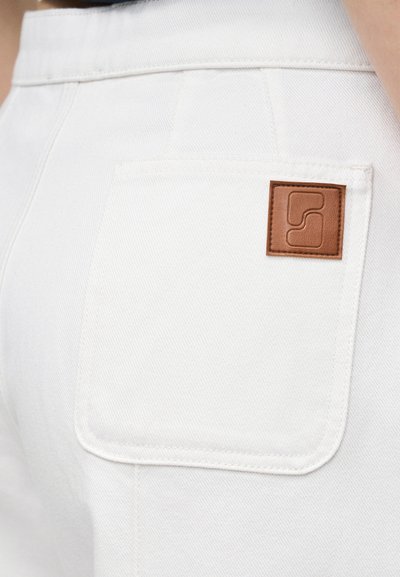 Πίσω τσέπη σε λευκό τζιν παντελόνι με ένα μικρό καφέ δερμάτινο έμπλαστρο που φέρει ένα γεωμετρικό ανάγλυφο λογότυπο.