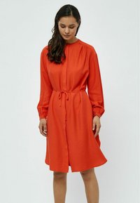 Punainen napillinen mekko, jossa on olkapäiden laskokset, pitkähihaiset hihat ja kangasvyö. Materiaali on sileä tekstuuriltaan ja rento siluetti.