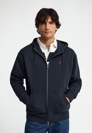 Joven con sudadera azul marino oscuro con cremallera y las manos en los bolsillos, sobre una camisa blanca con cuello, de pie frente a un fondo blanco liso.