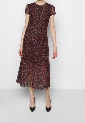 Femme portant une robe midi noire avec un motif floral rouge, des manches courtes et un tissu transparent, associée à des talons noirs à bout pointu.