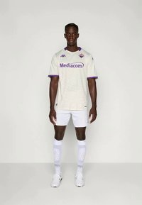 Camisa de fútbol en un tono crema claro, de tejido texturizado con acentos morados, que presenta el logo de Mediacom y un pequeño emblema. Combinada con pantalones cortos y calcetas blancas.