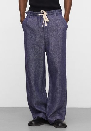 Pantaloni larghi in lino blu scuro con girovita beige con coulisse, indossati con scarpe nere e mani nelle tasche.