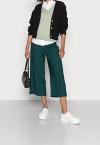 Cardigan noir côtelé porté sur un pull vert clair, chemise blanche à boutons, culottes vertes et baskets blanches à plateforme, tenant un sac noir.