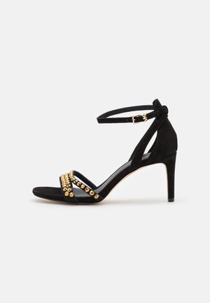 Schwarze High-Heel-Sandale mit Knöchelriemen und zwei vorderen Riemen, die mit goldenen runden Nieten auf weißem Hintergrund verziert sind.