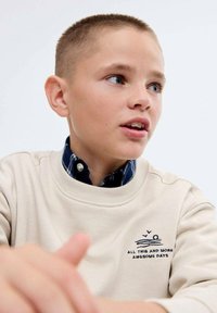 Sweat-shirt beige à col rond, brodé d'un soleil et du texte "TOUT ÇA ET ENCORE PLUS DE JOURNÉES GÉNIALES," superposé à une chemise à carreaux bleu marine et blanche.