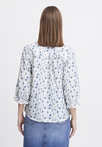 Blouse florale blanche avec des motifs bleus, manches bouffantes avec des poignets volants, coupe droite et détails de yoke au dos. Matière en coton.