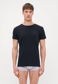 Schwarzes Baumwoll-T-Shirt mit rundem Ausschnitt und kurzen Ärmeln; auf der linken Brust befindet sich ein kleines Logo. Getragen mit hellgrauer Unterwäsche.