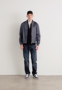 Chaqueta bomber gris sobre un sudadera negra, combinada con jeans azules y zapatillas negras/blancas. El modelo lleva una gorra negra.