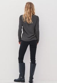 Woman with shoulder-length wavy blonde hair wearing a dark long-sleeve top, black pants, and knee-high black boots standing facing a white wall.