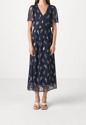 Frau trägt ein marineblaues Midikleid mit goldenen Blattmustern, transparenten kurzen Ärmeln und beigefarbenen Sandalen mit eckiger Spitze und Absatz.