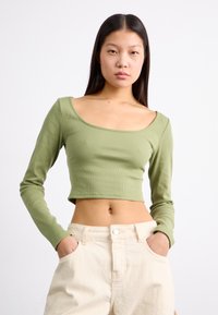 Crop top verde a maniche lunghe in tessuto a costine, con ampio scollo rotondo e silhouette aderente, abbinato a pantaloni alti color beige chiaro.
