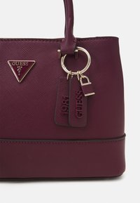Burgundy handbag in textured material featuring a gold-tone ring and tags with '1981' and 'GUESS' embossed. Includes a triangular logo plaque.