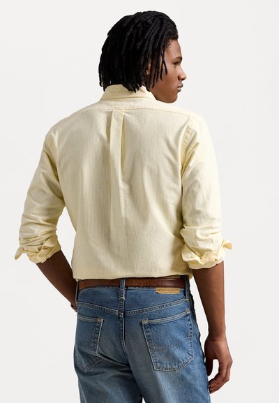 Camisa amarela clara, com riscas, de botão e mangas dobradas, combinada com jeans azuis. A camisa tem uma pala traseira reta e uma textura subtil.