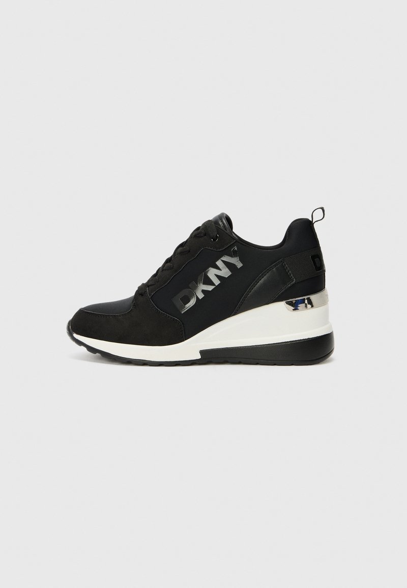 Black DKNY sneaker with a textured fabric upper, lace closure, silver logo accent, and a white wedge sole. Features a pull tab at the heel.
