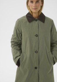 Olijfgroen lange jas met een bruine corduroy kraag, twee zijzakken en knoopsluitingen. Glad buitenoppervlak, middellange stijl.