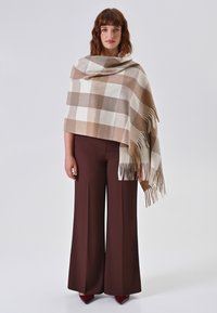 Bruine en crèmekleurige geblokte poncho met een zachte textuur, draperend ontwerp en franjes. Gecombineerd met brede bruine broek.