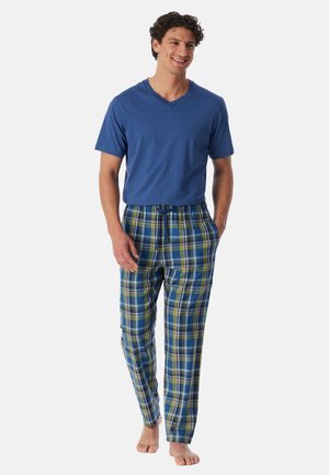 Smilende barfodet mand iført blå, kortærmet V-hals skjorte og blågrønternet pyjamasbukser med snorlukning, stående med hænderne i lommerne.