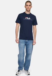 Camiseta de algodón azul marino de manga corta, con texto blanco y rojo de "FILA" y el año de establecimiento. Combinada con jeans de color azul claro y zapatillas.