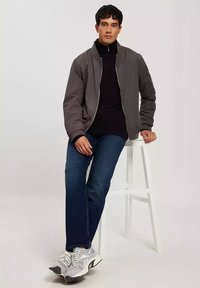 Szara kurtka bomberka na czarnej golfie, niebieskie dżinsy i szare sneakersy. Stoi na białym stołku, trzymając ręce na kolanach.