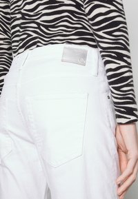 Personne portant un jean en denim blanc et un haut à manches longues rayé noir et blanc zébré, la main reposant le long du corps.