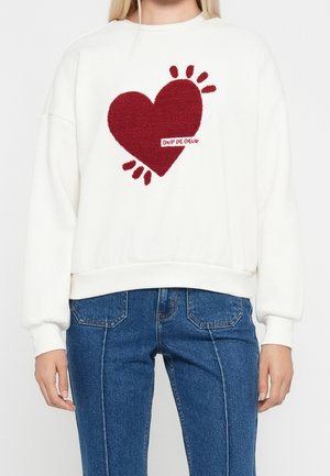 Sudadera blanca con un gran corazón rojo texturizado y el texto "COUP DE COEUR", combinada con pantalones vaqueros azules.