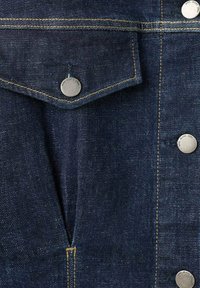 Chaqueta de denim azul oscuro con un bolsillo con tapa abotonado, botones plateados, costuras amarillas en contraste y una sutil textura granulada.