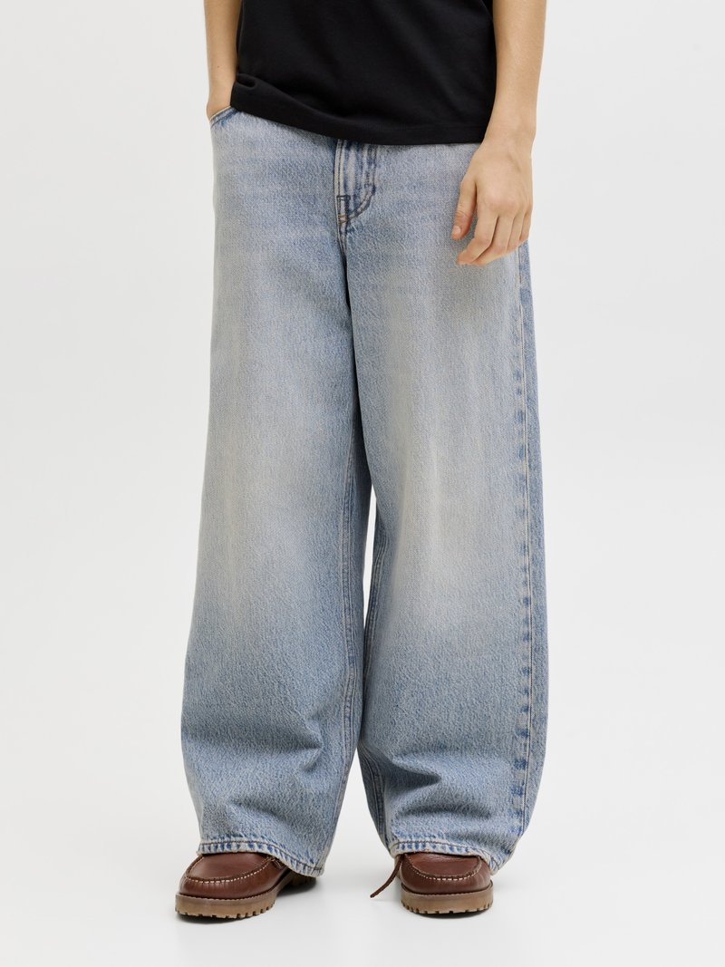 Persona vistiendo jeans azules desteñidos de pierna ancha con las manos parcialmente en los bolsillos y zapatos de cuero marrón contra un fondo liso.
