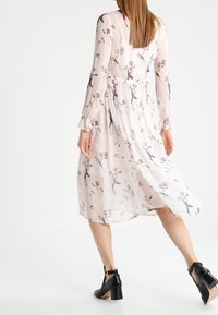 Floral print, lightweight, sheer fabric dress in pastel pink with long sleeves and a belted waist, paired with black heeled sandals.