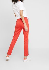 Červené sportovní kalhoty s bílými bočními pruhy, elastickým pasem, zužujícími se nohavicemi, v kombinaci s bílými teniskami. Hladký texturovaný materiál.