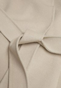 Detail van een beige wollen jas met een gestrikte riem, een gladde textuur en een gestructureerde revers. Benadrukt zachte stof en nauwkeurige afwerking.