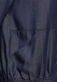 Marineblaue Jacke mit glatter Textur, ausgestattet mit einer Fronttasche und einem gerafften Saum. Leichter Stoff mit subtiler glänzender Oberfläche.