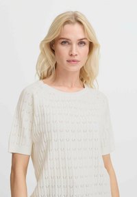 Lichtbeige gebreide top met korte mouwen, voorzien van een ribbeltextuur en kleine gaatjespatronen door de stof.